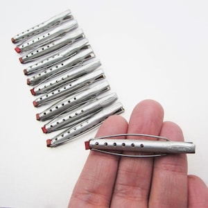 May include: A collection of vintage silver-toned hair rollers with red rubber ends. One roller is held in a hand, while the others are arranged on a white surface. The rollers have a series of small holes along their length.