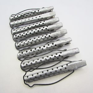 May include: A collection of eight silver-colored hair rollers with a perforated design. Each roller has a series of small holes and a black wire clip. The rollers are arranged on a white surface.