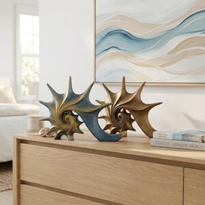 May include: Two sculptural seashell figurines displayed on a wooden surface. One shell features a gradient of metallic blue and gold, while the other is a solid metallic gold. Both shells have a spiraled center and pointed, radiating edges.