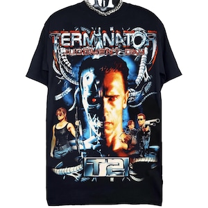 Peut inclure: Un t-shirt noir à manches courtes avec une grande impression graphique du film Terminator 2 : Le Jugement dernier. Le motif comprend le titre du film, un visage de cyborg central et des personnages armés, entourés de tentacules mécaniques.