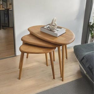 May include: A set of three nested tables made of light-toned wood with smooth, rounded, oval-shaped tops and tapered legs. A rectangular book with a white cover and a small, abstract white sculpture are placed on the largest table surface.