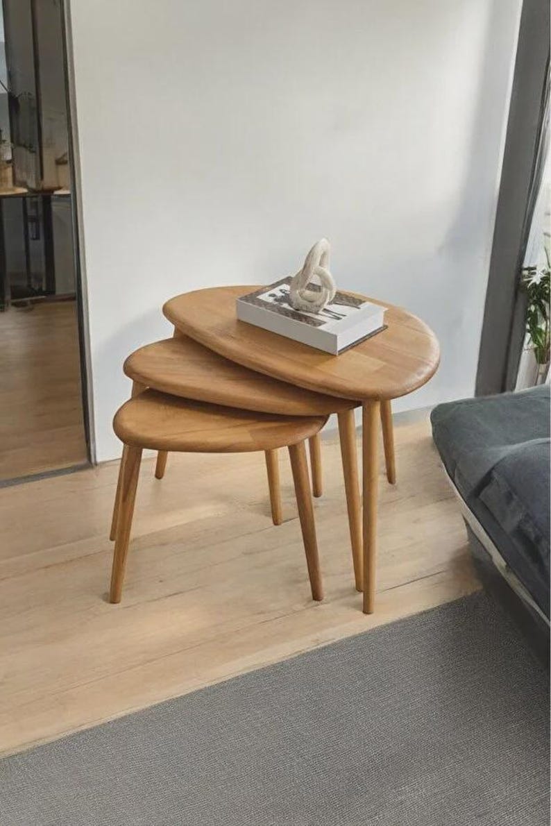 May include: A set of three nested tables made of light-toned wood with smooth, rounded, oval-shaped tops and tapered legs. A rectangular book with a white cover and a small, abstract white sculpture are placed on the largest table surface.
