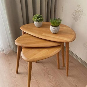 May include: A set of three nesting tables made of light-colored wood with smooth, rounded, pebble-shaped tops and tapered legs. Two small potted artificial plants in textured white containers sit on the largest table surface.
