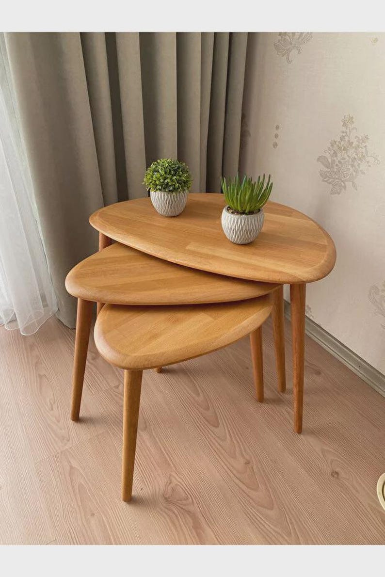 May include: A set of three nesting tables made of light-colored wood with smooth, rounded, pebble-shaped tops and tapered legs. Two small potted artificial plants in textured white containers sit on the largest table surface.