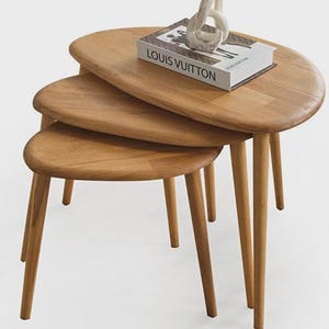 May include: A set of three light brown wooden nesting tables with rounded, organic-shaped tops and tapered legs. A white book with the text LOUIS VUITTON printed in black sits on the largest table, topped with a small, off-white abstract sculpture.