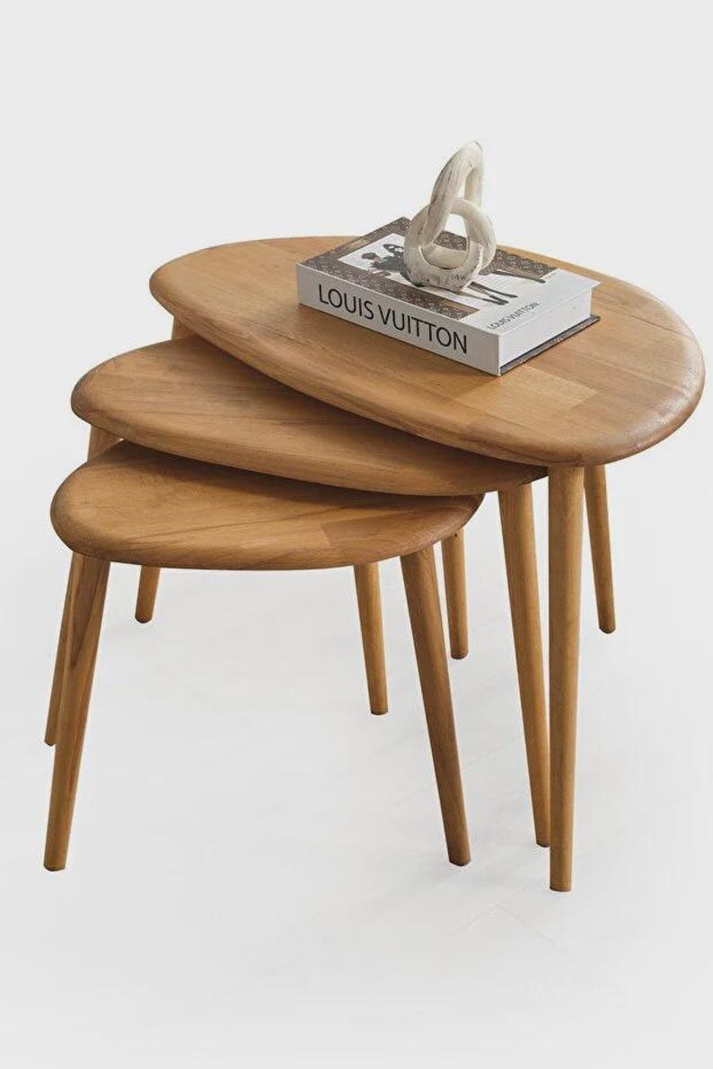 May include: A set of three light brown wooden nesting tables with rounded, organic-shaped tops and tapered legs. A white book with the text LOUIS VUITTON printed in black sits on the largest table, topped with a small, off-white abstract sculpture.