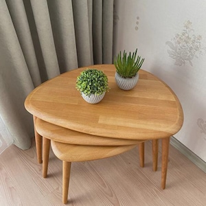 May include: A set of three light-toned wooden nesting tables with rounded triangular tops and tapered legs. The tables are stacked in a tiered arrangement, with two small potted green plants placed on the surface of the largest top table.