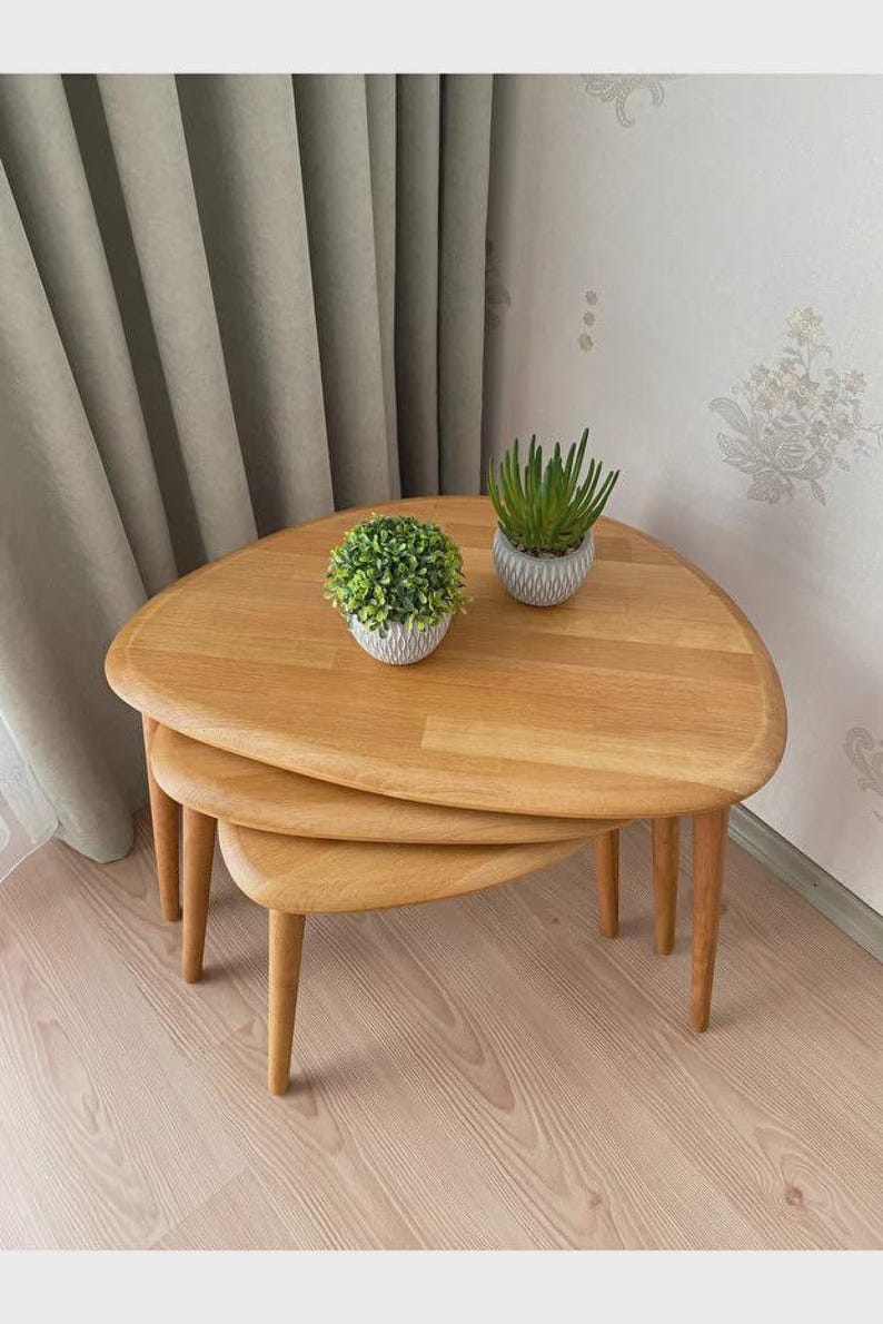 May include: A set of three light-toned wooden nesting tables with rounded triangular tops and tapered legs. The tables are stacked in a tiered arrangement, with two small potted green plants placed on the surface of the largest top table.