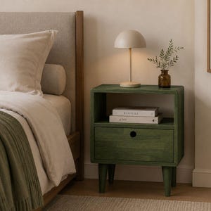 May include: A dark green wooden nightstand featuring an open storage shelf and a single drawer with a circular cutout handle. The nightstand stands on four tapered legs and is positioned next to a bed with a beige headboard and green throw blanket.
