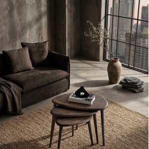 May include: A set of three nesting coffee tables with dark brown wooden tops and tapered legs. The tables are arranged in a tiered stack, with a black abstract sculpture and a book titled Simplicity placed on the top surface of the largest table.