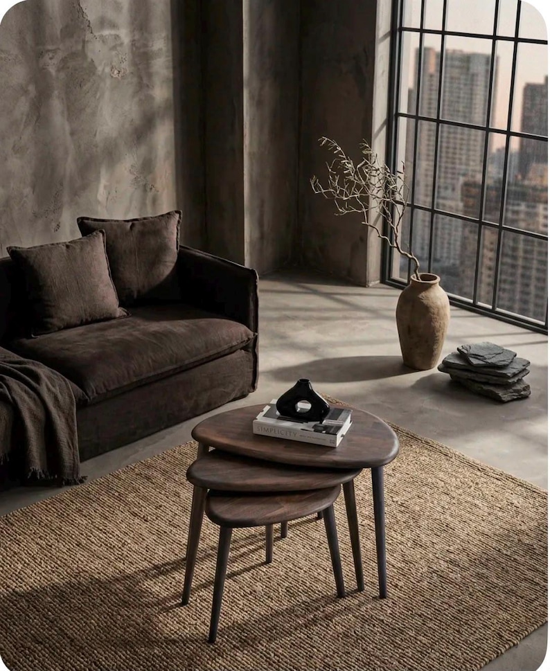May include: A set of three nesting coffee tables with dark brown wooden tops and tapered legs. The tables are arranged in a tiered stack, with a black abstract sculpture and a book titled Simplicity placed on the top surface of the largest table.