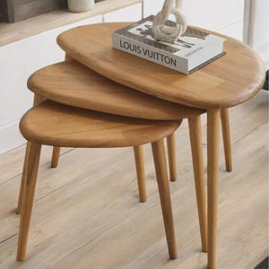 May include: A set of three light brown wooden nesting tables with rounded, oval-shaped tops and tapered legs. A white book with the text LOUIS VUITTON printed in black and a white abstract sculptural object are placed on the largest table.