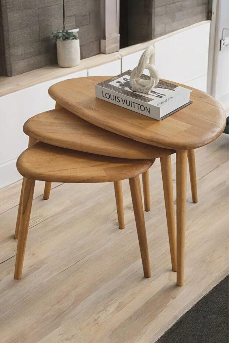 May include: A set of three light brown wooden nesting tables with rounded, oval-shaped tops and tapered legs. A white book with the text LOUIS VUITTON printed in black and a white abstract sculptural object are placed on the largest table.