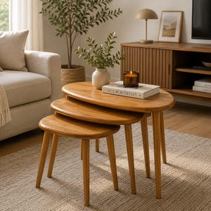 May include: A set of three nesting tables crafted from light-toned wood, each featuring an oval-shaped tabletop and four tapered legs. The tables are arranged in a cascading formation, with the largest table supporting a white vase and a book.