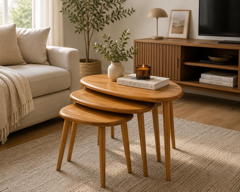 May include: A set of three nesting tables crafted from light-toned wood, each featuring an oval-shaped tabletop and four tapered legs. The tables are arranged in a cascading formation, with the largest table supporting a white vase and a book.