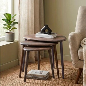 May include: A set of three nesting tables with dark brown wooden oval tops and tapered legs. A black abstract ceramic vase sits on a book titled Simplicity on the top table. Two additional books titled At Home and The Quiet Hours are stacked below.