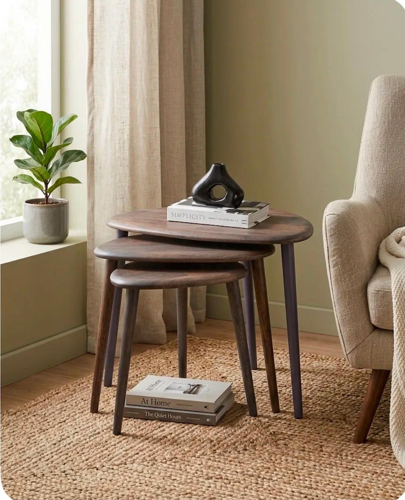 May include: A set of three nesting tables with dark brown wooden oval tops and tapered legs. A black abstract ceramic vase sits on a book titled Simplicity on the top table. Two additional books titled At Home and The Quiet Hours are stacked below.