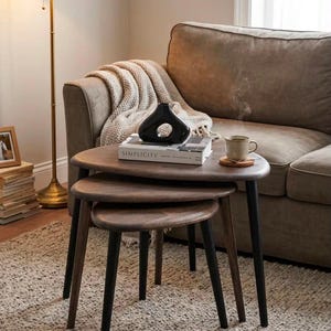 May include: A set of three nesting tables with dark brown wooden tops and black legs. The tables are arranged in a tiered formation. A white book titled SIMPLICITY, a black abstract vase, and a steaming ceramic mug sit on the largest table.