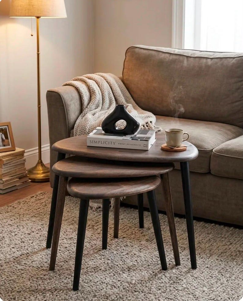 May include: A set of three nesting tables with dark brown wooden tops and black legs. The tables are arranged in a tiered formation. A white book titled SIMPLICITY, a black abstract vase, and a steaming ceramic mug sit on the largest table.