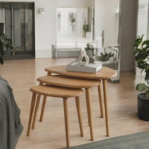 May include: A set of three nesting tables made of light-toned wood, each featuring a smooth, rounded triangular top and three tapered legs. The tables are arranged in a descending height order, with a white decorative object resting on top.
