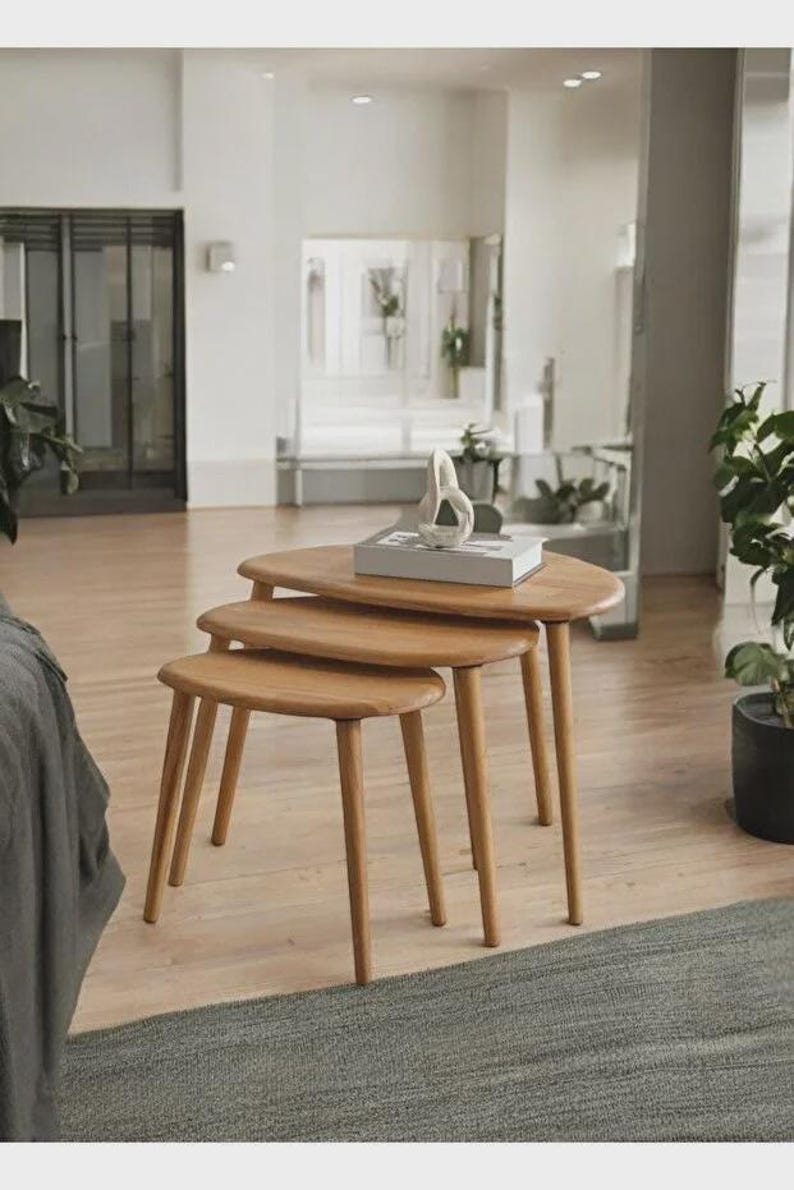 May include: A set of three nesting tables made of light-toned wood, each featuring a smooth, rounded triangular top and three tapered legs. The tables are arranged in a descending height order, with a white decorative object resting on top.