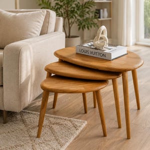 May include: A set of three nesting tables crafted from light-toned wood, featuring smooth, rounded, oval-shaped tabletops and tapered legs. A white book with the text LOUIS VUITTON and a white abstract sculptural object sit on the largest table.