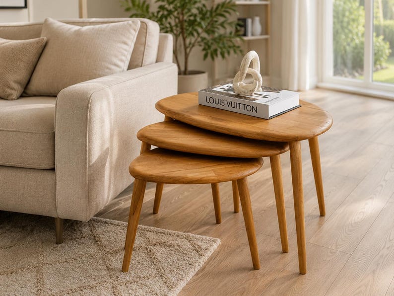 May include: A set of three nesting tables crafted from light-toned wood, featuring smooth, rounded, oval-shaped tabletops and tapered legs. A white book with the text LOUIS VUITTON and a white abstract sculptural object sit on the largest table.
