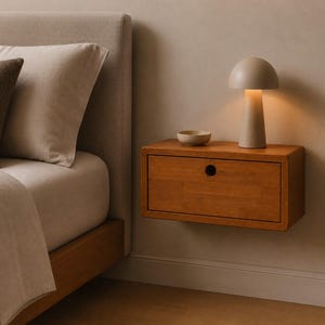 May include: A rectangular wooden floating nightstand with a single drawer featuring a circular cutout handle. A small beige bowl and a mushroom-shaped table lamp with a warm glowing light are placed on the flat top surface of the nightstand.