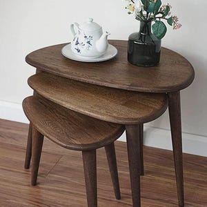 May include: A set of three nesting tables crafted from dark brown wood with a visible grain, featuring rounded triangular tops and tapered legs. A white teapot with blue floral patterns and a dark green glass vase with white flowers sit on top.