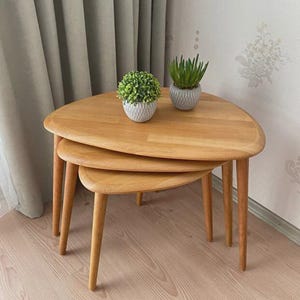 May include: A set of three light-toned wooden nesting tables with rounded triangular tops and tapered legs. Two small potted artificial green plants in textured white and gray containers are placed on the largest table surface.