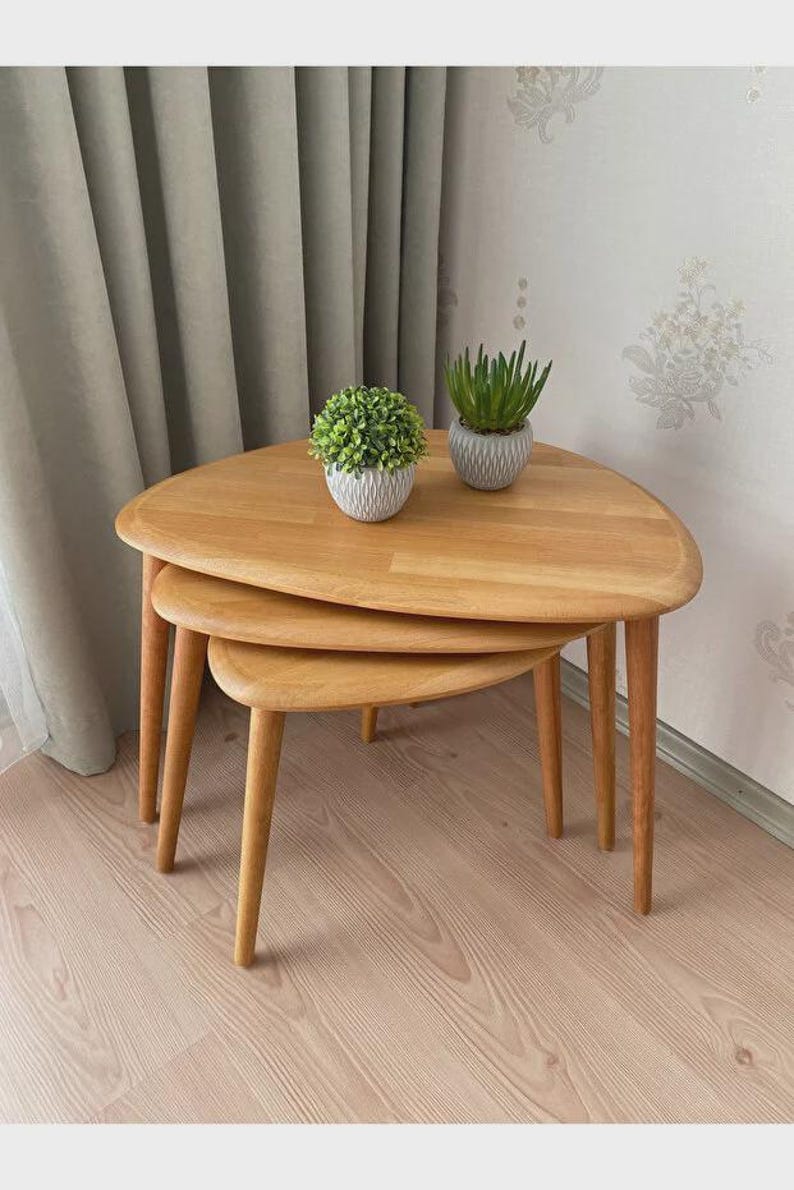May include: A set of three light-toned wooden nesting tables with rounded triangular tops and tapered legs. Two small potted artificial green plants in textured white and gray containers are placed on the largest table surface.