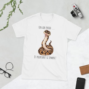 May include: A white t-shirt featuring a graphic of a brown and tan patterned snake with large eyes and a pink tongue. The text above the snake reads "UN AIR FROID ..." and the text below the snake reads "ET POURTANT SI SYMPA !" in black font.