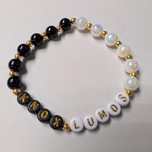 May include: A beaded bracelet featuring a mix of black, white, and iridescent beads separated by small gold-tone spacers. The bracelet includes black letter beads spelling KNOX and white letter beads spelling LUMOS in gold-colored lettering.