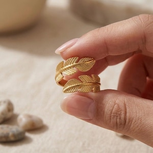 Peut inclure: Une bague dorée au design enveloppant ornée de deux motifs de plumes détaillés. La surface métallique présente une finition texturée qui souligne le motif des nervures des feuilles, formant un anneau continu autour du doigt.