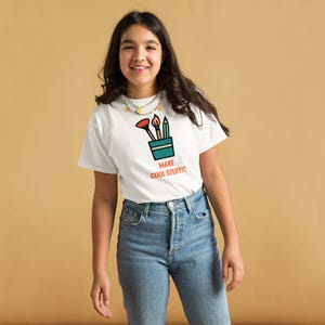 Può includere: Una t-shirt bianca a maniche corte con la grafica di una tazza color verde acqua contenente un pennello rosso, un pennello marrone e una matita verde acqua. Sotto la grafica, il testo "MAKE COOL STUFF!!!" è scritto in lettere maiuscole arancioni.