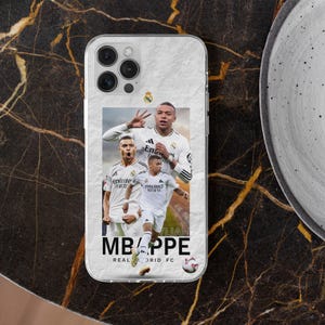 Puede incluir: Una funda transparente para smartphone con un gr&aacute;fico de tres jugadores de f&uacute;tbol con uniformes blancos del Real Madrid. El dise&ntilde;o incluye el texto MBAPPE y REAL MADRID FC, junto con un peque&ntilde;o icono de bal&oacute;n de f&uacute;tbol y el escudo del club.
