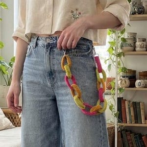 May include: A beige button-down shirt with a small floral embroidery, light blue wide-leg denim jeans with a raw hem, and a multi-colored crocheted chain link accessory attached to the belt loop, paired with white canvas low-top sneakers.