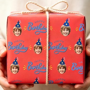 May include: A square gift box wrapped in red paper featuring a repeating pattern of a smiling child wearing a blue party hat with yellow stars and the text Happy Birthday in blue script, tied with a thin beige twine bow on top.