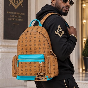 Könnte beinhalten: Ein hellbrauner Rucksack mit wiederkehrendem Logo-Muster, leuchtend blauen Akzenten und einer Vordertasche mit einem hängenden rechteckigen Anhänger, der den Text FAITH HOPE CHARITY trägt. Der Rucksack hat einen Tragegriff, zwei Seitentaschen und goldfarbene Reißverschlüsse.