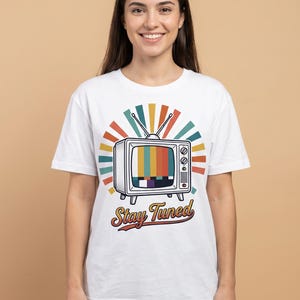 Könnte beinhalten: Ein weißes Kurzarm-T-Shirt mit einer Grafik eines Vintage-Fernsehers mit Antenne und einem farbigen Testbildschirm. Hinter dem Fernseher befindet sich ein Strahlenmuster in gedeckten Farben. Der Text Stay Tuned ist darunter aufgedruckt.