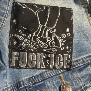 May include: A light blue denim jacket featuring a square black fabric patch sewn onto the chest. The patch displays a white graphic of a foot crushing ice shards and the text FUCK ICE in bold, blocky, white capital letters.
