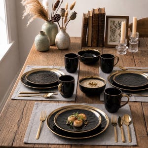 Op de afbeelding: Een servies bestaande uit zwarte borden en kommen met gouden randen, vergezeld van goudkleurig bestek, zwarte mokken met gouden oren en grijze stoffen placemats, gerangschikt op een houten tafel met decoratieve vazen en kaarsen.