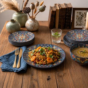 Puede incluir: Un juego de platos redondos de cerámica y un cuenco con base azul oscuro y complejos patrones florales en naranja, amarillo y verde azulado. La vajilla está dispuesta sobre una mesa de madera con una servilleta azul y cubiertos dorados.