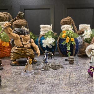 May include: A collection of crocheted potion bottles with various colors and embellishments, including flowers, leaves, and a small wooden barrel.  A miniature figure of a warrior stands next to a miniature figure of a knight.  There are also several dice on the table.