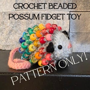 Może przedstawiać: Szydełkowa zabawka typu fidget z oposa z tęczowymi koralikami. Zabawka ma szary korpus, różowy ogon oraz czarne i różowe rysy twarzy. Tekst na zdjęciu brzmi "CROCHET BEADED POSSUM FIDGET TOY" i "PATTERN ONLY!"