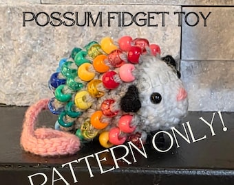 May include: A crocheted possum fidget toy with rainbow-colored beaded accents. The toy has a gray body, pink tail, and black and pink facial features. Text on the image reads "CROCHET BEADED POSSUM FIDGET TOY" and "PATTERN ONLY!"