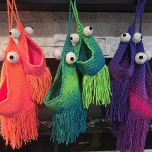 May include: Three hanging, handmade crochet monster bags in vibrant colors. The bags are orange, green, and purple, each with large white and black googly eyes and a fringe of yarn at the bottom. The bags are suspended from a string.