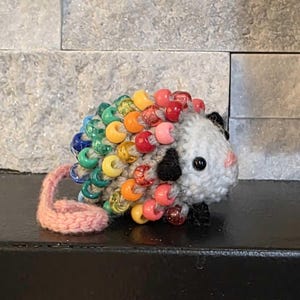 May include: A handmade, gray possum figurine with a rainbow beaded back. The possum has a pink tail, black ears, and a black spot on its face. The beads are in shades of blue, green, yellow, orange, and pink.