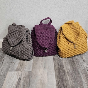 May include: Three crocheted mini backpacks in gray, purple, and yellow. Each backpack has a small, round, gold-colored zipper pull.