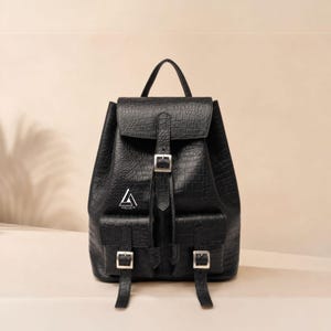 Puede incluir: Una mochila negra con textura de piel de cocodrilo, con asa superior, solapa principal con cierre de hebilla plateada y dos bolsillos frontales con hebillas plateadas. Un logotipo triangular blanco con el texto ASTRA LEATHER está al frente.